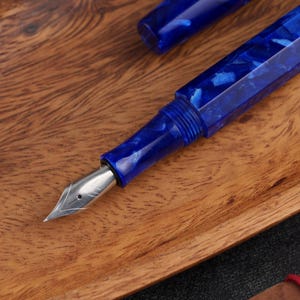 May include: A close-up of a blue fountain pen with a silver nib, set against a wooden surface. The pen's body has a marbled design with shades of blue. The cap is visible in the background.