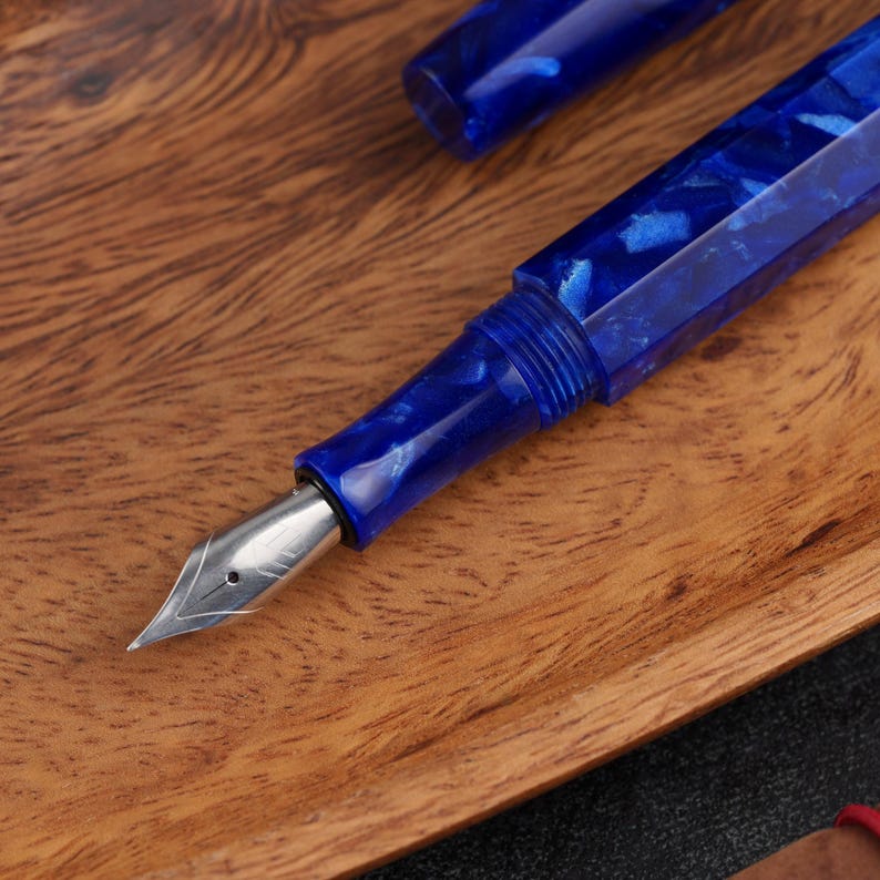 May include: A close-up of a blue fountain pen with a silver nib, set against a wooden surface. The pen's body has a marbled design with shades of blue. The cap is visible in the background.