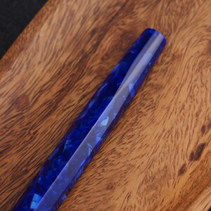 May include: A faceted, blue resin pen body with a marbled appearance. The pen is displayed on a wooden surface with a natural grain pattern. The pen's design features a deep blue color with lighter blue and white inclusions.