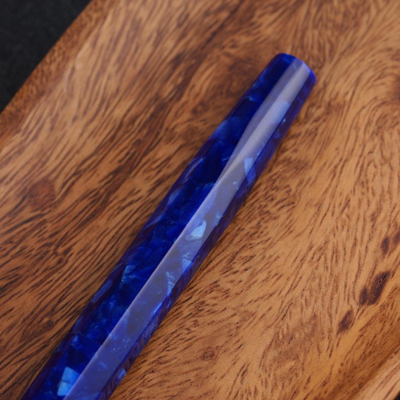 May include: A faceted, blue resin pen body with a marbled appearance. The pen is displayed on a wooden surface with a natural grain pattern. The pen's design features a deep blue color with lighter blue and white inclusions.