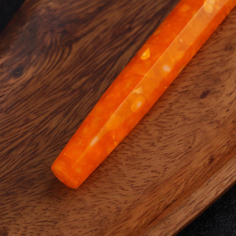 May include: Close-up of an orange and white pen. The pen has a faceted design and a glossy finish. The pen is resting on a wooden surface with a natural wood grain pattern. The pen's color is a vibrant orange with white marbling.