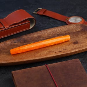 May include: An orange faceted pen rests on a wooden tray, alongside a brown leather watch with a gold buckle, a leather pen case, and a brown leather journal. The pen has a translucent, speckled appearance.