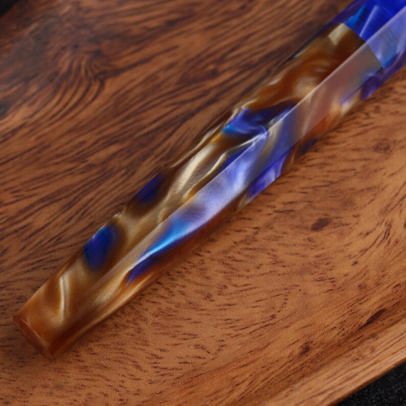 May include: A cylindrical pen body featuring a marbled pattern of swirling blue, metallic gold, and brown resin. The surface has a faceted, multi-sided design with smooth, polished edges that catch the light, creating a reflective appearance.