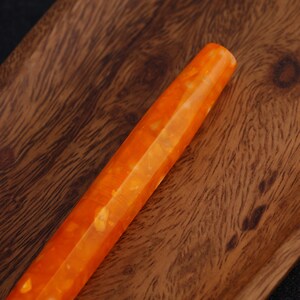 May include: Close-up of an orange and yellow pen with a faceted design. The pen has a marbled appearance and is set against a wooden background. The pen's body is a vibrant orange color with yellow marbling.