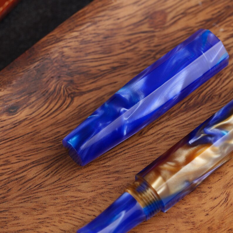 May include: A fountain pen cap and body featuring a marbled blue and white resin pattern with a faceted, multi-sided design. The pen body includes a threaded section in a metallic gold tone, showcasing a swirling, polished finish throughout.