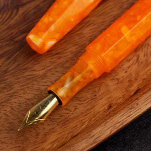 May include: Close-up of an orange fountain pen with a gold nib. The pen's body has a marbled appearance, and the cap is detached, resting beside the pen. The pen is set on a wooden surface.