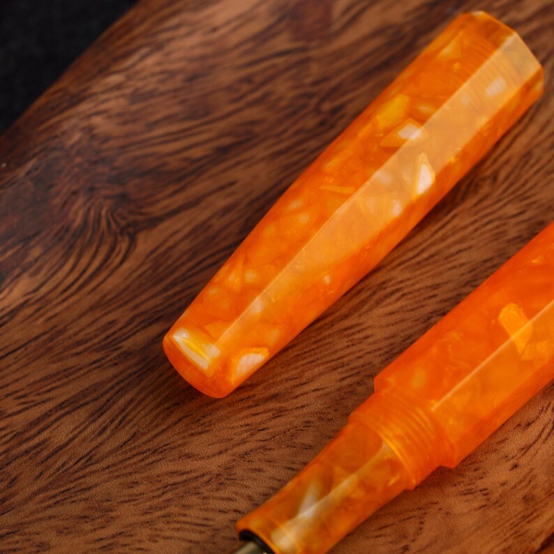 May include: Close-up of two orange and white marbled fountain pen parts. The pen parts are set against a dark wood grain background. The pen parts have a faceted design and a glossy finish.