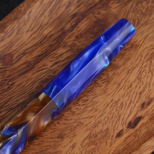 May include: A cylindrical writing instrument barrel featuring a polished resin finish with swirling patterns of deep blue, translucent white, and metallic bronze tones. The material has a glossy, reflective surface with a smooth, tapered shape.