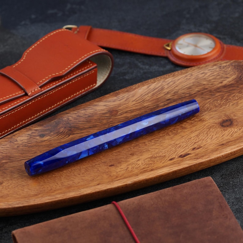 May include: A faceted, blue resin pen with a marbled pattern rests on a wooden tray. A brown leather pen case and a watch with a brown leather strap are in the background. The pen is a striking writing instrument.