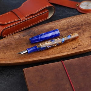 May include: A fountain pen with a marbled blue and cream resin body and a matching blue cap. The pen features a silver-colored metal nib and a blue grip section. The cap is detached and lies horizontally next to the pen body on a wooden surface.