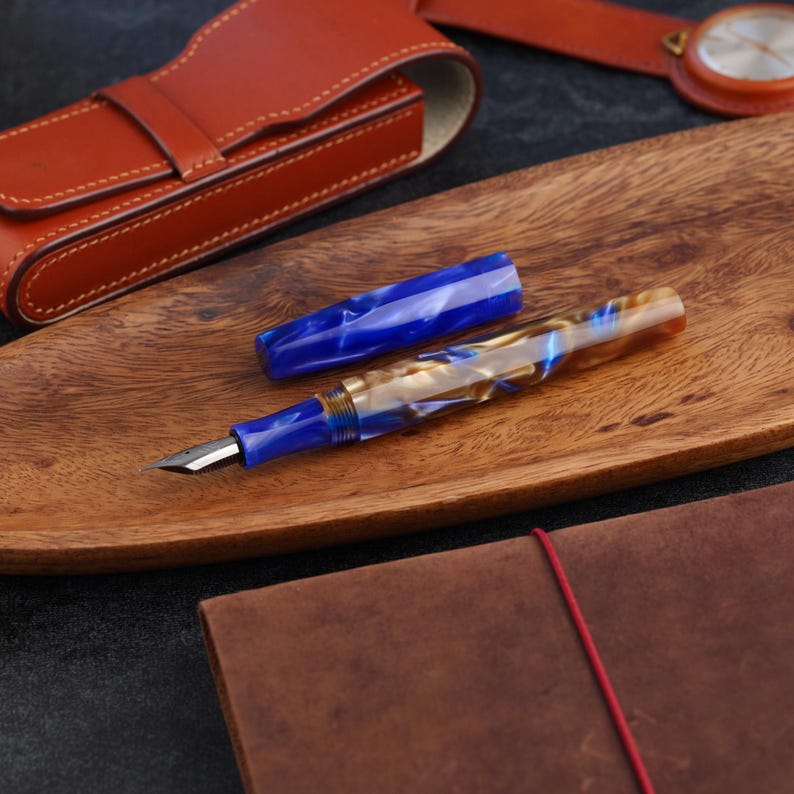 May include: A fountain pen with a marbled blue and cream resin body and a matching blue cap. The pen features a silver-colored metal nib and a blue grip section. The cap is detached and lies horizontally next to the pen body on a wooden surface.