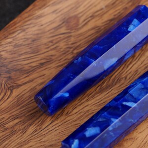 May include: Close-up of two faceted, blue resin pen blanks with a marbled appearance. The blanks are a deep, saturated blue with lighter blue and white inclusions, set against a warm, wooden surface.