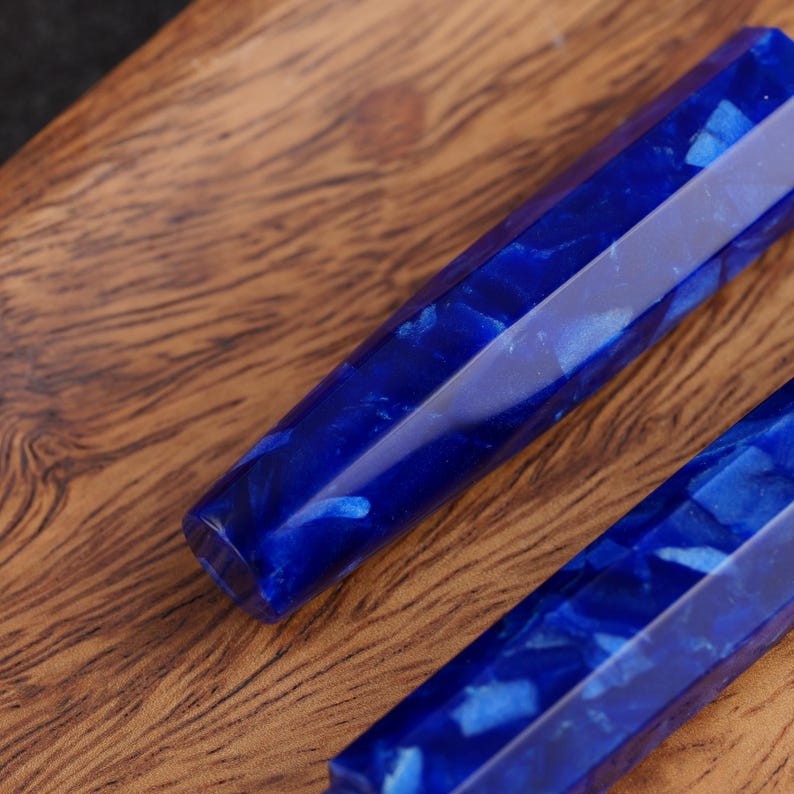 May include: Close-up of two faceted, blue resin pen blanks with a marbled appearance. The blanks are a deep, saturated blue with lighter blue and white inclusions, set against a warm, wooden surface.