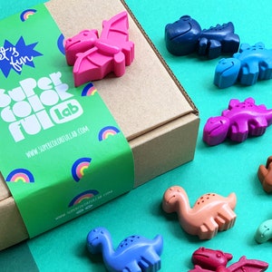 Puede incluir: Una caja de crayones con forma de dinosaurio en una variedad de colores, incluyendo rosa, azul, verde, naranja y rojo. La caja está etiquetada como "Super Color Fun Lab" y tiene un diseño de arcoíris. El texto "Let's Fun" también está en la caja.