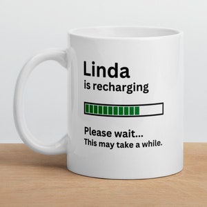 Może przedstawiać: Biały kubek ceramiczny z czarno-zielonym wzorem. Na kubku widnieje napis "Linda is recharging" z paskiem postępu i tekstem "Please wait... This may take a while."