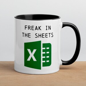 Peut inclure: Mug en céramique blanche avec intérieur et anse noirs. Le mug affiche le texte "FREAK IN THE SHEETS" au-dessus d'un logo Excel vert. Le mug est posé sur une surface en bois.