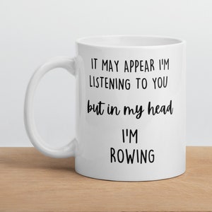 May include: White ceramic coffee mug with black text that reads "It may appear I'm listening to you but in my head I'm rowing".