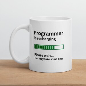Könnte beinhalten: Eine weiße Keramiktasse mit einem schwarzen und grünen Design. Die Tasse trägt den Schriftzug "Programmer is recharging" mit einer Fortschrittsanzeige und dem Text "Please wait... This may take some time."