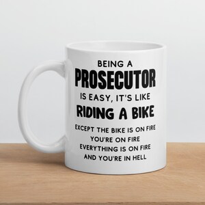 May include: White ceramic coffee mug with black text that reads "Being a Prosecutor is easy, it's like riding a bike except the bike is on fire, you're on fire, everything is on fire and you're in hell."