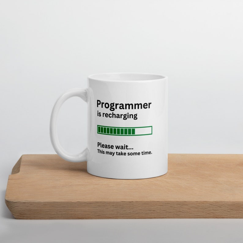 Peut inclure: Une tasse en c&eacute;ramique blanche avec un motif noir et vert. La tasse affiche "Programmer is recharging" avec une barre de progression et le texte "Please wait... This may take some time."