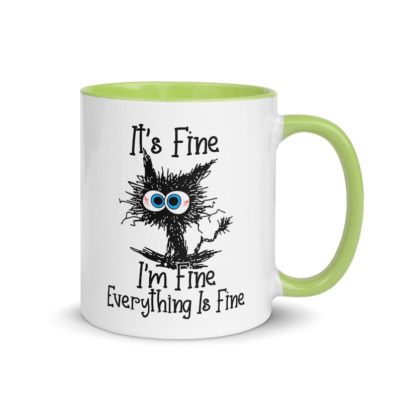 May include: White ceramic mug with a green rim and handle. The mug features a black and white illustration of a cat with the text "It's Fine I'm Fine Everything Is Fine."