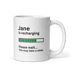 Op de afbeelding: Een witte keramische mok met een zwart en groen ontwerp. De mok heeft de tekst "Jane is recharging" met een voortgangsbalk en de tekst "Please wait... This may take a while."