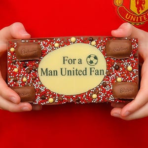Może przedstawiać: Prostokątna tabliczka czekolady z napisem "For a Man United Fan" i piłką nożną. Czekolada jest ozdobiona czerwonymi, białymi i brązowymi posypkami oraz czterema mniejszymi kawałkami czekolady. Tło to czerwona koszulka z logo Manchester United.