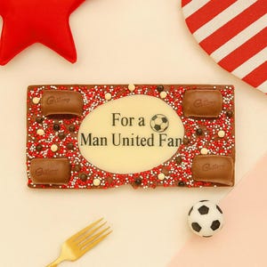 Może przedstawiać: Prostokątna tabliczka czekolady z napisem "For a Man United Fan" i grafiką piłki nożnej. Tabliczka jest ozdobiona czerwono-białymi posypkami, kawałkami czekolady i czterema kwadratami czekolady Cadbury. Obecna jest r&oacute;wnież mała piłka nożna.
