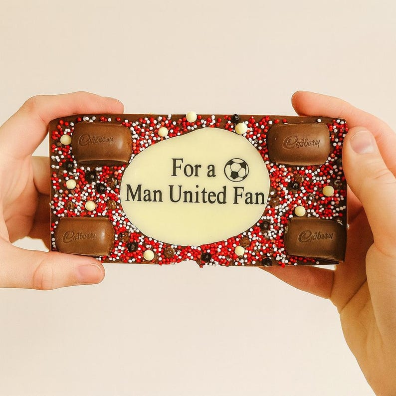 Może przedstawiać: Prostokątna czekolada Cadbury z białym owalem pośrodku z napisem "For a Man United Fan" z piłką nożną. Czekolada jest ozdobiona czerwonymi, białymi i czarnymi posypkami oraz czterema kawałkami czekolady Cadbury.