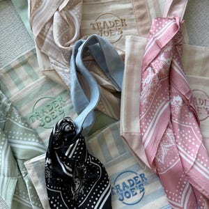 May include: A collection of canvas tote bags featuring the Trader Joe's logo, each paired with a coordinating patterned scarf. The set includes bags and scarves in shades of beige, sage green, light blue, and black, with pink and white accents.