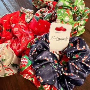 Könnte beinhalten: Ein Set mit 6 Weihnachts-Scrunchies in Rot, Grün, Weiß und Blau mit verschiedenen Mustern, darunter Zuckerstangen, Schneeflocken und Weihnachtsbäume. Die Scrunchies sind aus Satin gefertigt und werden mit einem weißen Etikett mit der Aufschrift "Elegant Wrangler Boutique" geliefert.