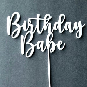Op de afbeelding: Een witte taarttopper met de woorden "Birthday Babe" in een cursief schrift.