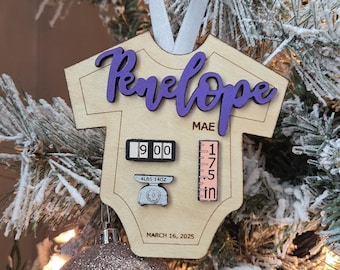 May include: A light-coloured wooden ornament shaped like a baby onesie, featuring the purple name Penelope Mae, a clock showing 9:00, a scale reading 4lbs 14oz, a ruler marked 17.5 in, and the date MARCH 16, 2025, all suspended by a white ribbon.