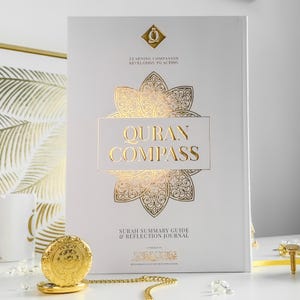 May include: A white book titled "Quran Compass" with gold foil lettering and a decorative floral design. The book is a "Surah Summary Guide & Reflection Journal." A gold pocket watch and chain are in the foreground.