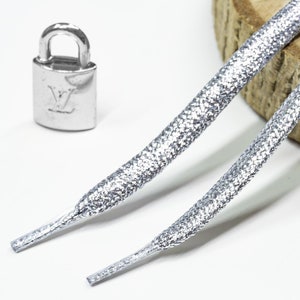 Puede incluir: Dos cordones de zapatos plateados con un dije de candado plateado. Los cordones tienen un acabado texturizado brillante.