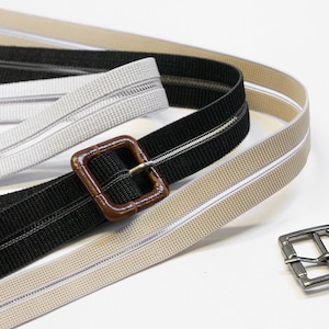 Può includere: Tre tipi diversi di fettuccia elastica con una fibbia marrone. La fettuccia è beige, bianca e nera.