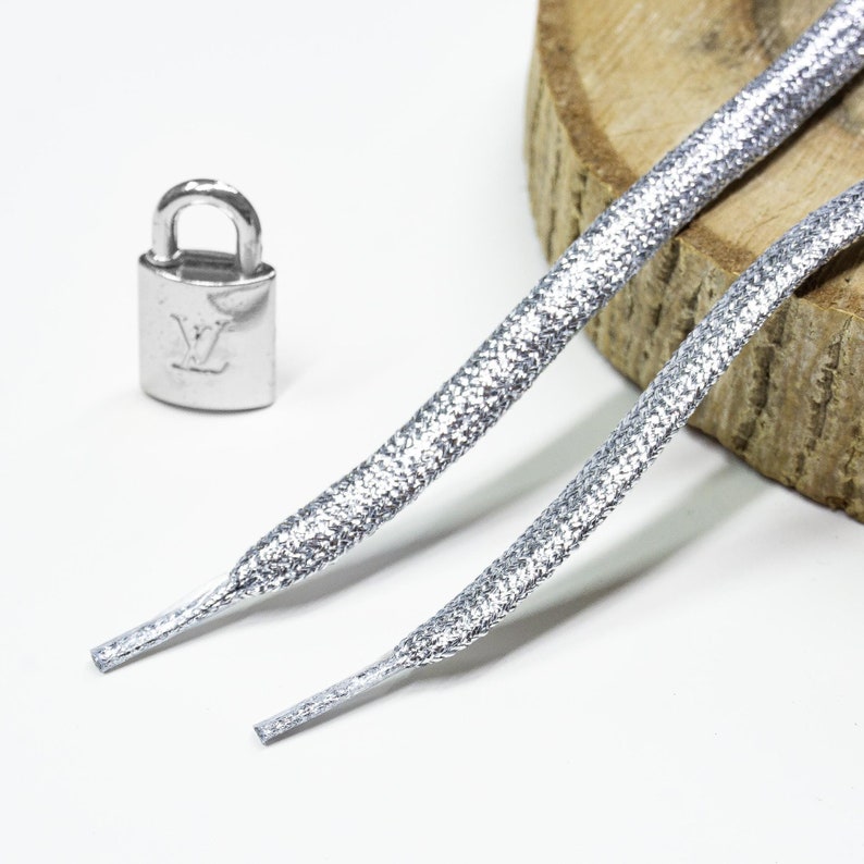 Puede incluir: Dos cordones de zapatos plateados con un dije de candado plateado. Los cordones tienen un acabado texturizado brillante.