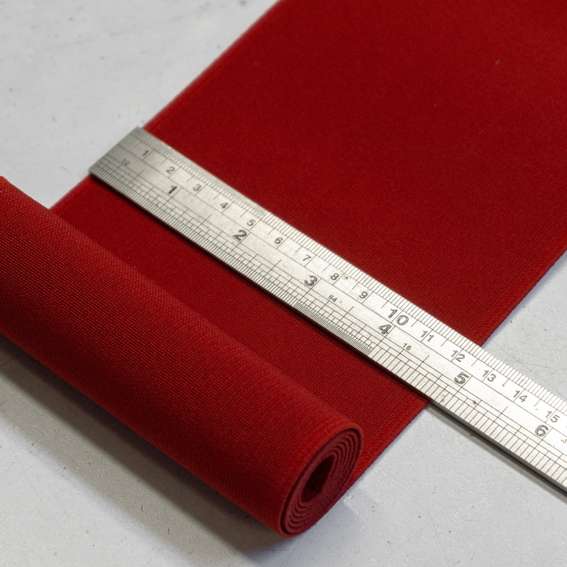 May include: A red fabric ribbon is rolled up and measured with a ruler. The ruler is marked in inches and shows a measurement of 15 inches.