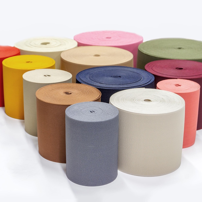 May include: Assortment of colourful fabric rolls, including red, yellow, beige, brown, blue, green, and pink. The rolls are arranged in a semi-circle on a white surface.