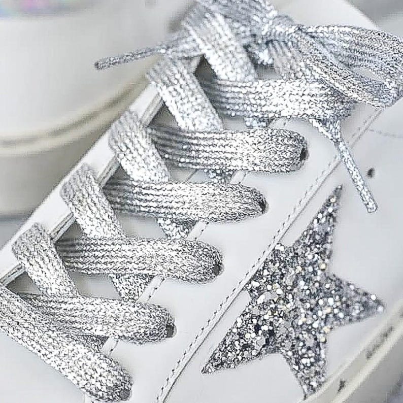 Puede incluir: Zapatillas blancas de cuero con detalle de estrella de purpurina plateada y cordones met&aacute;licos plateados.