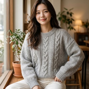 May include: A light gray long-sleeve sweater featuring a prominent vertical cable knit pattern on the front, a ribbed crew neckline, and ribbed cuffs. The garment is constructed from a textured knit fabric with a soft, heathered appearance.