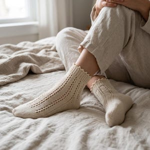 May include: A pair of cream-colored knit socks featuring a delicate openwork pattern along the foot and ankle, finished with a scalloped ribbed cuff. The socks are crafted with a visible vertical stitch design and a reinforced heel section.