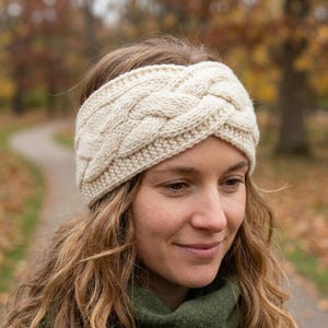 May include: A cream-colored knit headband featuring a prominent central cable-knit braid pattern. The accessory is constructed with a thick, textured yarn and includes a ribbed border along the top and bottom edges, providing a snug fit.