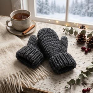 Peut inclure: Une paire de moufles tricotées gris foncé avec un poignet côtelé épais et un motif de mailles texturées. Les moufles sont fabriquées à partir d'un fil épais et sont placées ensemble, mettant en valeur la construction régulière et la couleur anthracite.
