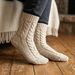 May include: A pair of beige cable-knit socks featuring a vertical braided pattern and a ribbed cuff. The socks are crafted from a textured yarn and extend to the mid-calf, showcasing a classic knit construction with a reinforced heel and toe.