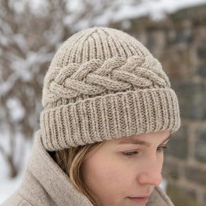May include: A beige knit beanie featuring a prominent cable knit braid pattern encircling the crown, finished with a ribbed cuff at the base. The hat is constructed with a thick, textured yarn in a neutral tan color, showcasing a classic design.