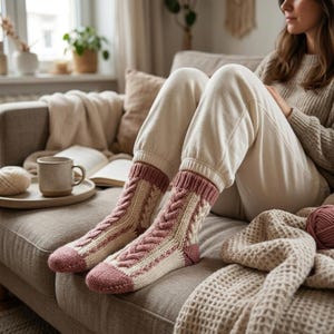 May include: A pair of knit socks featuring a two-tone design with cream-colored panels and dusty rose accents. The socks display a prominent vertical cable knit pattern running up the center, finished with ribbed cuffs and solid-colored toes.