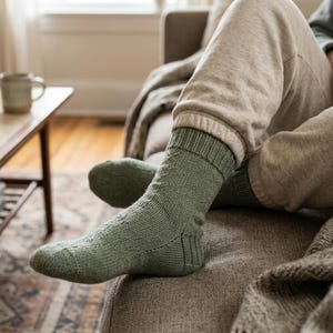 Peut inclure: Une paire de chaussettes en tricot vert sauge avec un revers côtelé et un motif torsadé discret sur le côté. Les chaussettes présentent une texture tricotée visible et montent jusqu'au milieu du mollet, avec un talon et une pointe renforcés.