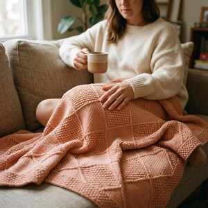 Könnte beinhalten: Eine weiche, pfirsichfarbene Strickdecke mit einem strukturierten Rautenmuster aus abwechselnden rechten und linken Maschen. Die Decke liegt über einem Schoß und zeigt die dicke Garnstruktur und das geometrische Design.