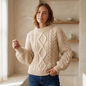 Puede incluir: Un suéter de punto trenzado en color beige con un patrón de diamantes prominente en la parte delantera, un cuello redondo grueso acanalado y mangas largas con puños acanalados. La textura incluye trenzas verticales y punto arroz.